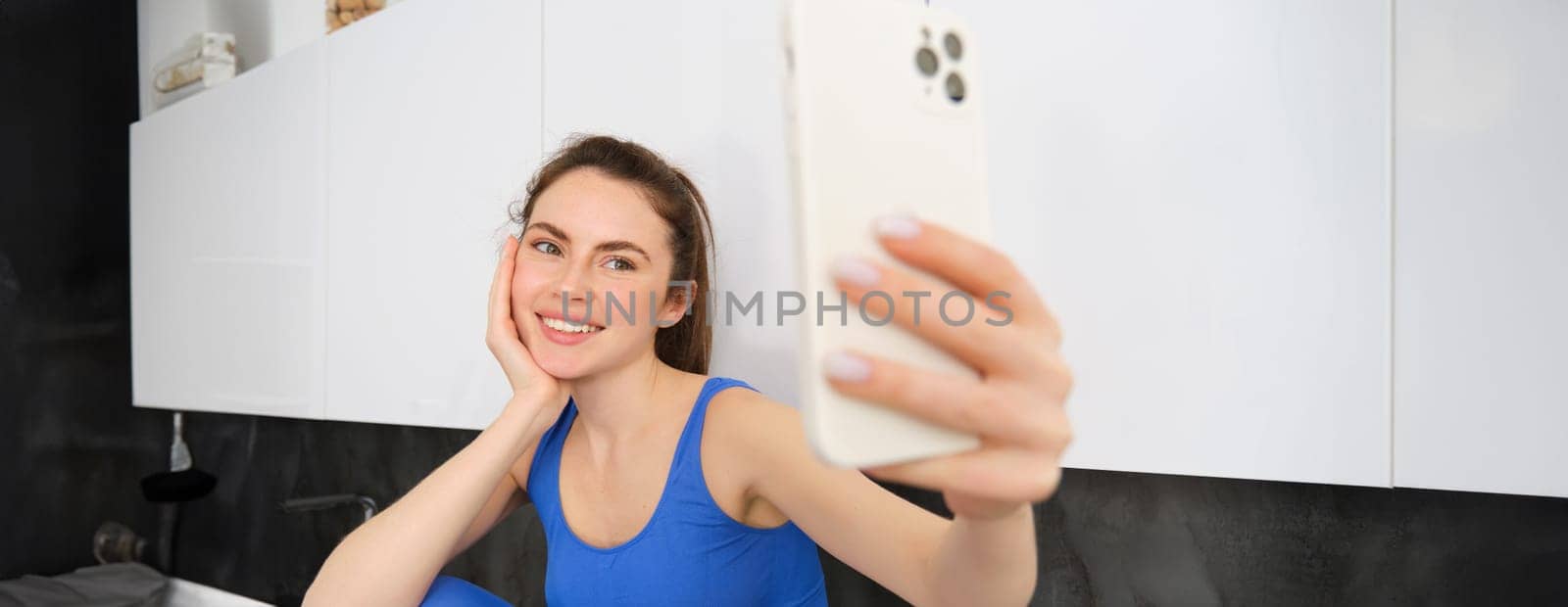Portrait of fitness girl posing for photo, taking selfie on smartphone app, sitting in kitchen, wearing activewear.