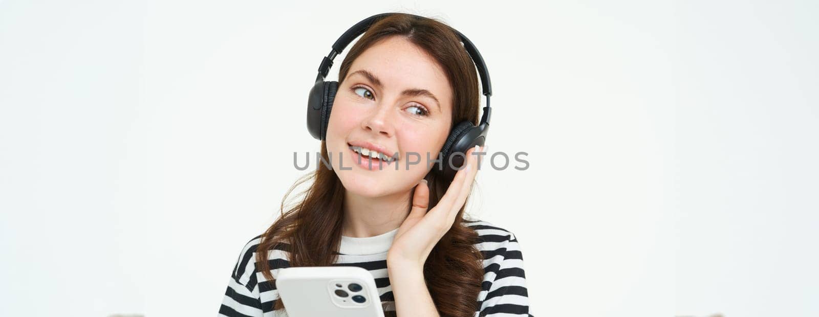 Smiling, beautiful female student in headphones, holding mobile phone, smiling and listening music, using smartphone, standing over white background.