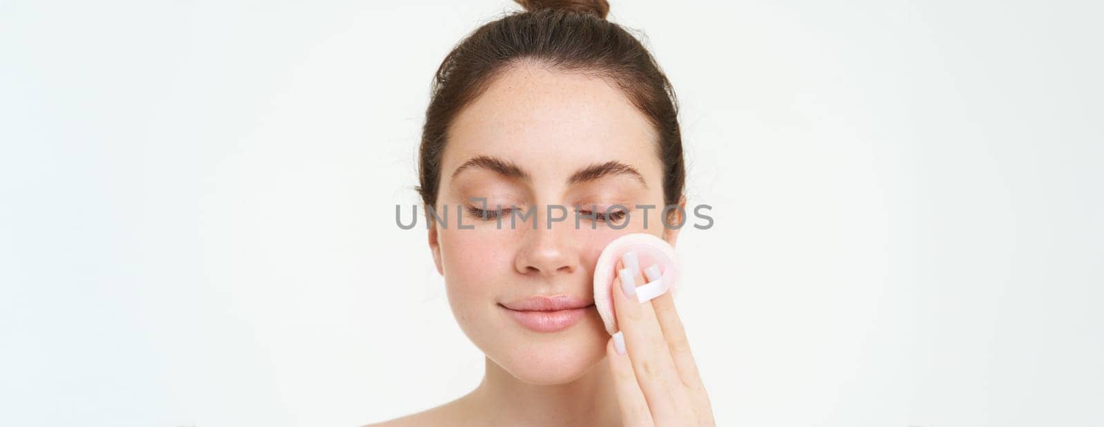 Image of young woman taking off her makeup with cotton pads, using facial cleanser, cleaning her face with skincare treatment, standing over white background.