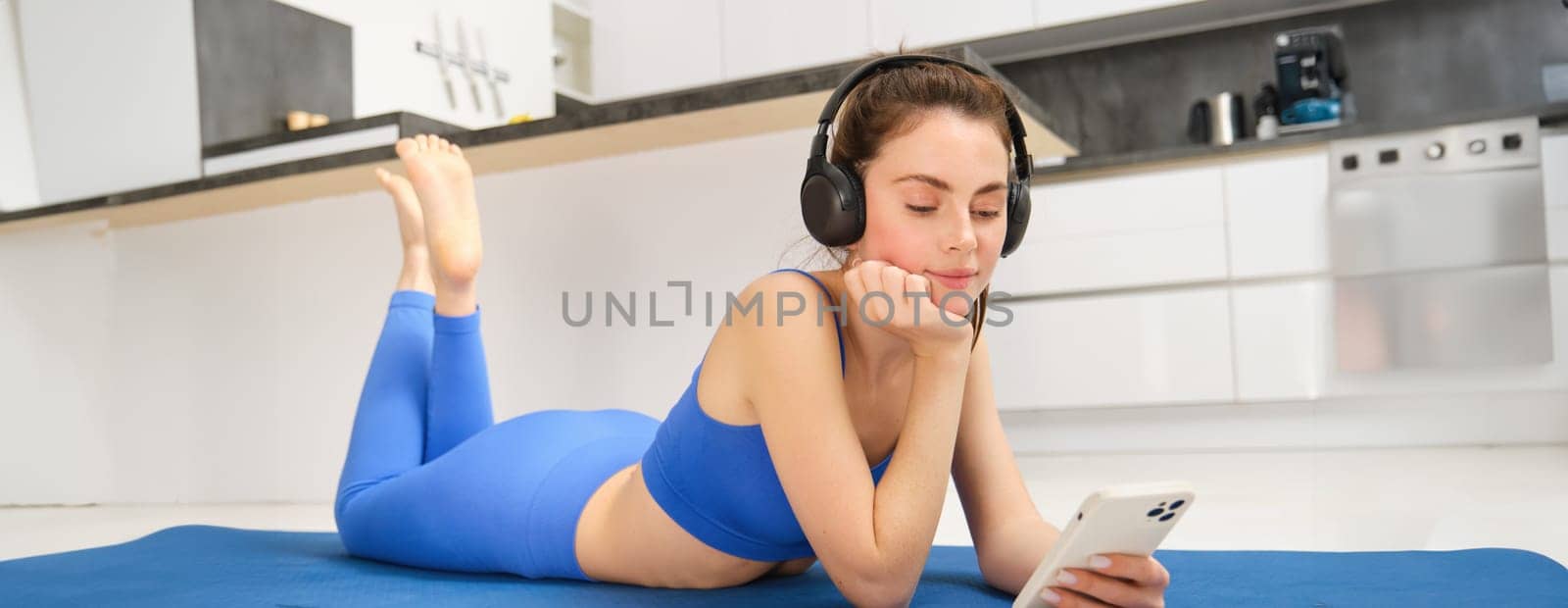 Portrait of brunette fitness girl, laying on yoga mat at home, watching workout tutorials, videos on smartphone, wearing sportswear and wireless headphones.