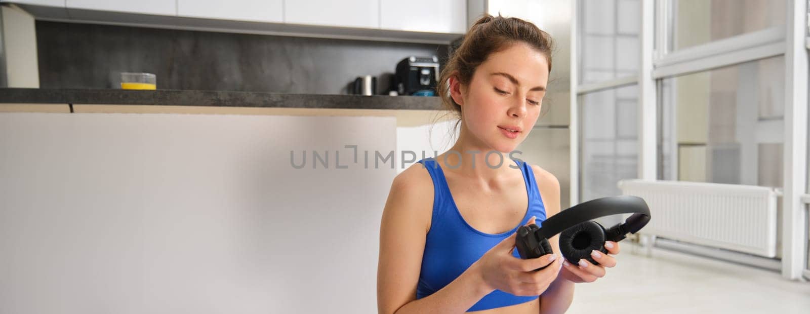 Image of young fitness girl, wearing blue tracksuit, puts on wireless headphones to workout with music, does yoga at home by Benzoix