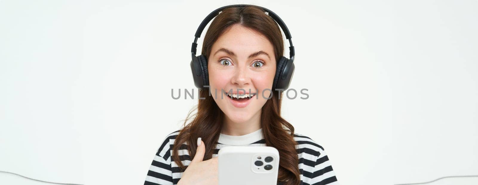 Young woman with surprised face, looking at smartphone screen, reacts amazed, looks with happiness at mobile phone, standing in wireless headphones, white background by Benzoix