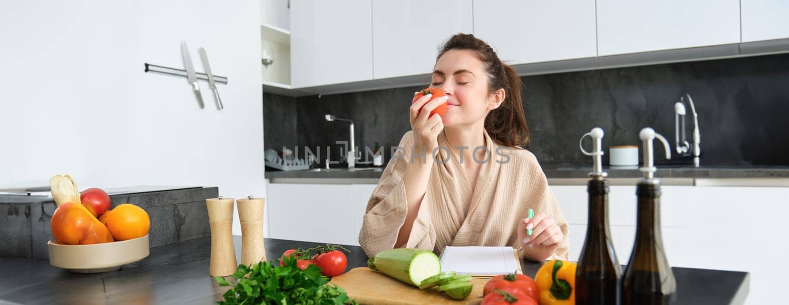 Portrait of beautiful young woman in the kitchen, writing down cooking recipe, sitting near chopping board with vegetables and making grocery list, creating healthy veggie menu for her family by Benzoix
