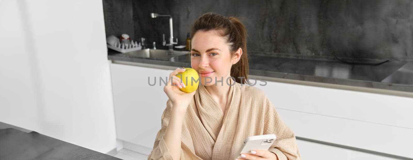 Image of attractive young woman in bathrobe, sits in kitchen, eats green apple and looks at mobile phone, uses smartphone app, orders food on application.