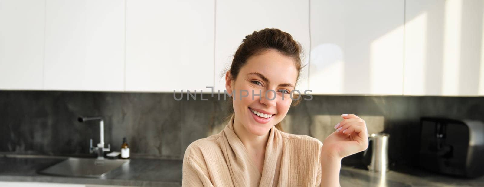 Portrait of beautiful and happy woman standing in kitchen in bathrobe, posing and smiling at camera, cooking food, preparing meal, eating lunch near counter.