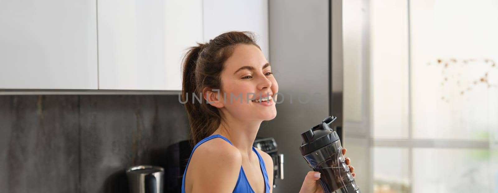Image of fit and healthy, fitness woman in sportswear, drinking water, holding bottle shaker and standing in kitchen, staying hydrated after workout.