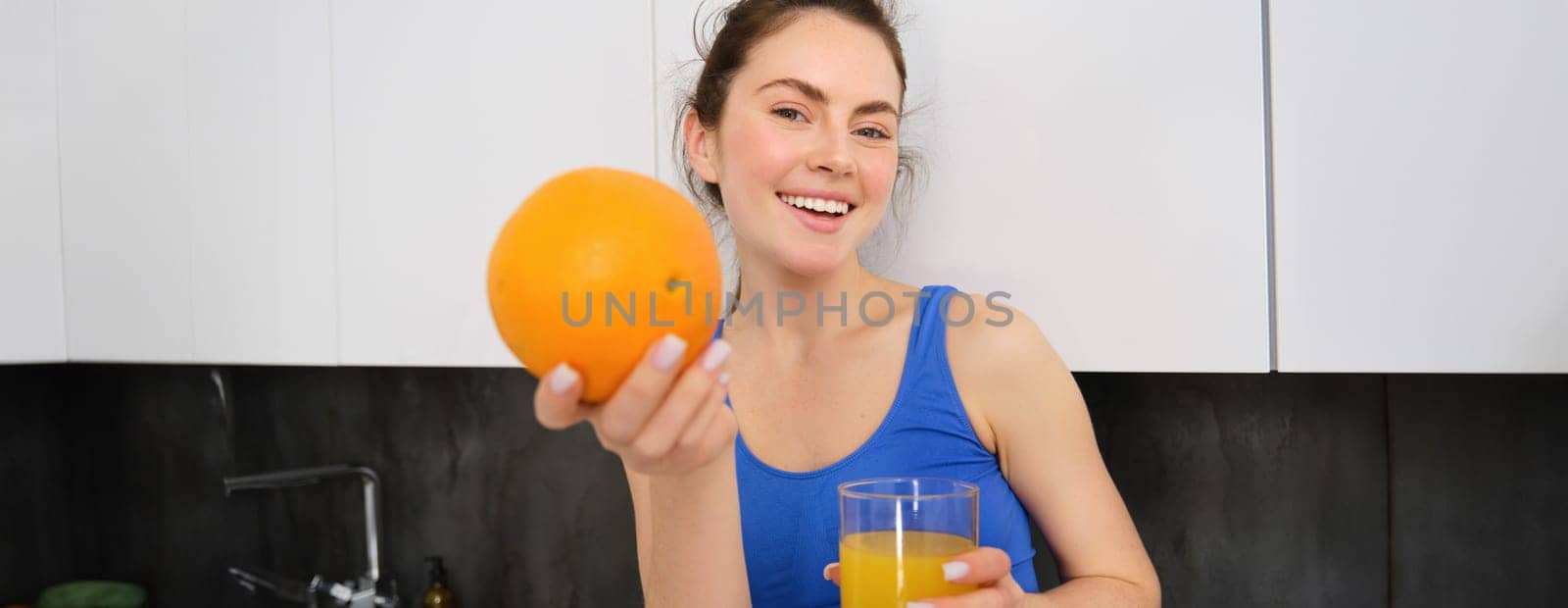 Close up portrait of sportswoman, fitness girl holding glass of fresh juice and an orange in hands, smiling at camera, standing in kitchen. Workout and healthy lifestyle concept