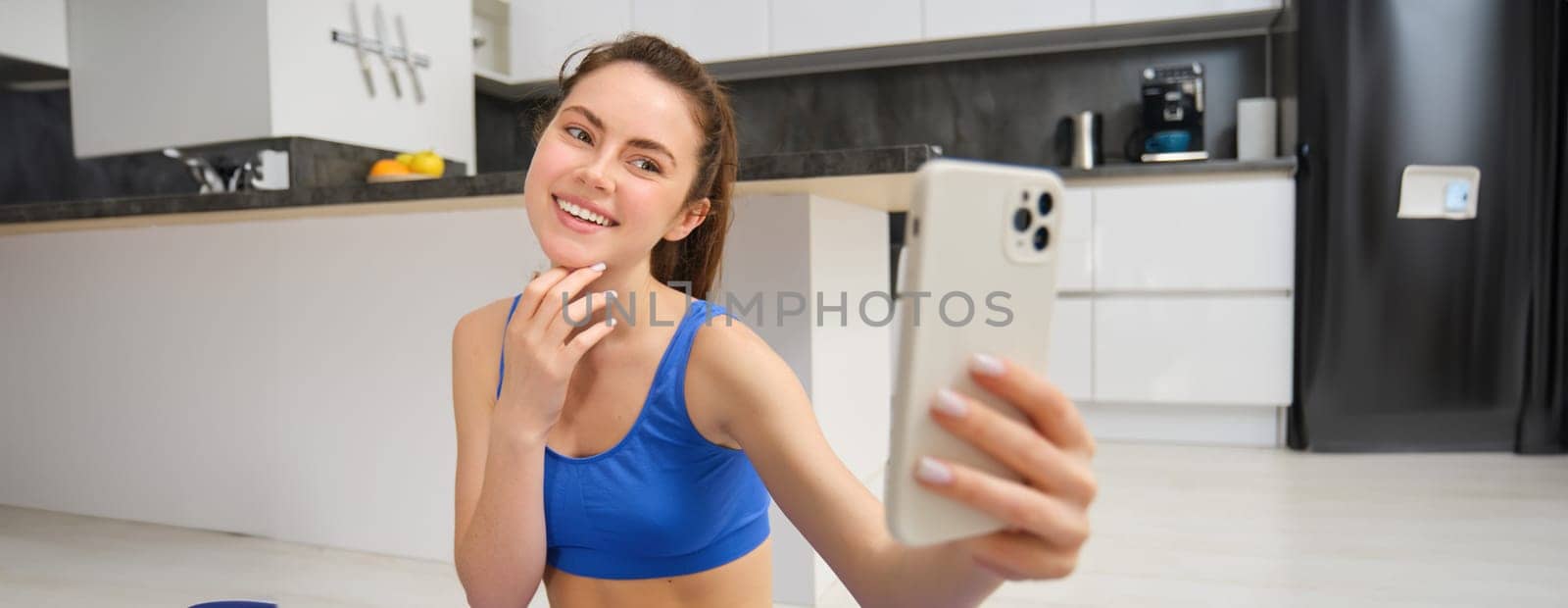 Portrait of young fitness girl, blogger records her workout from home, poses for selfie on smartphone, does yoga on camera, sits in blue sportsbra and leggings.