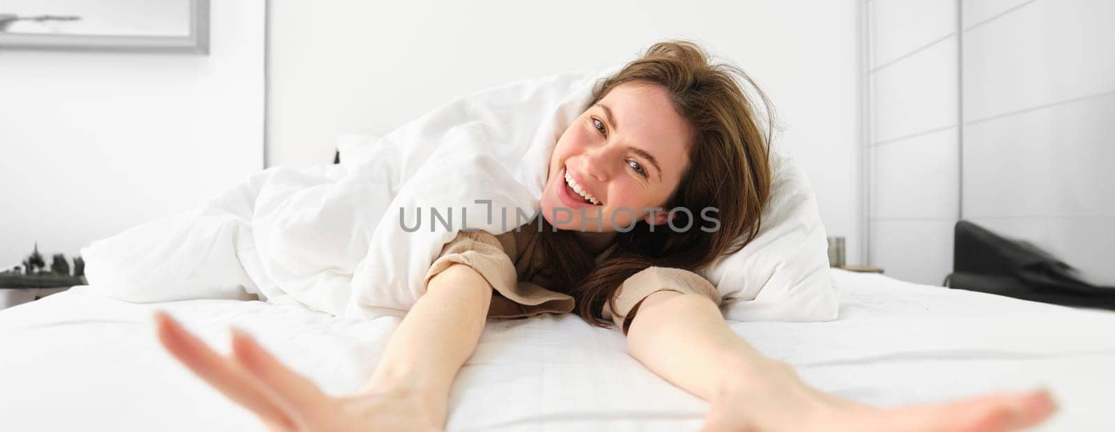 Portrait of happy young woman waking up energetic in morning, stretching arms towards camera and laughing in her bed under blankets.