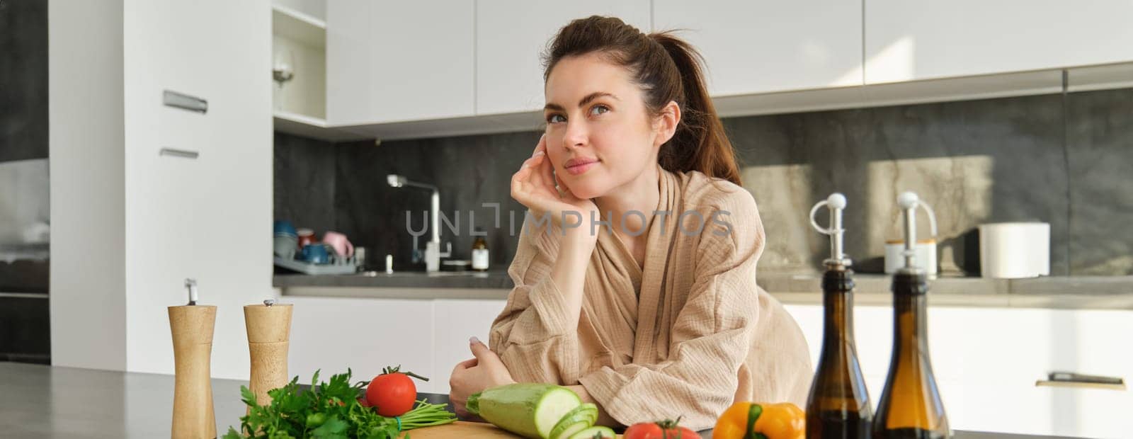 Portrait of pretty woman in bathrobe, standing in kitchen with thoughtful face, posing near vegetables and bottle of oil, thinking what food to prepare, making salad, eating healthy.