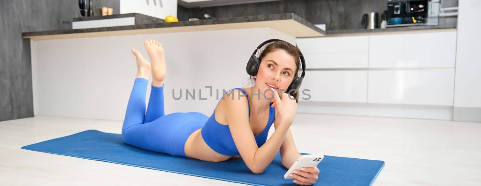 Image of smiling young woman in fitness clothing, listening music in wireless headphones, holding smartphone, using sports app while doing workout at home.