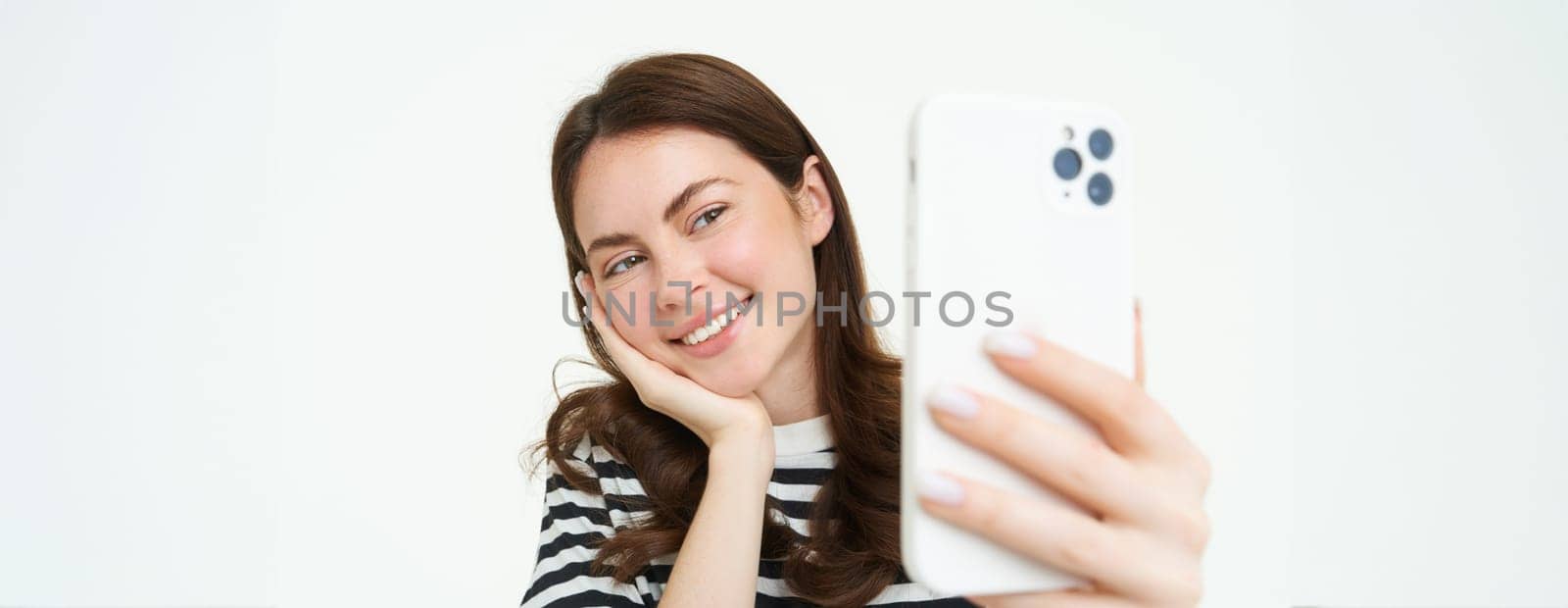 Portrait of young woman taking selfies on smartphone, posing for photo,. using mobile phone app for taking funny and cute pictures, isolated on white background.