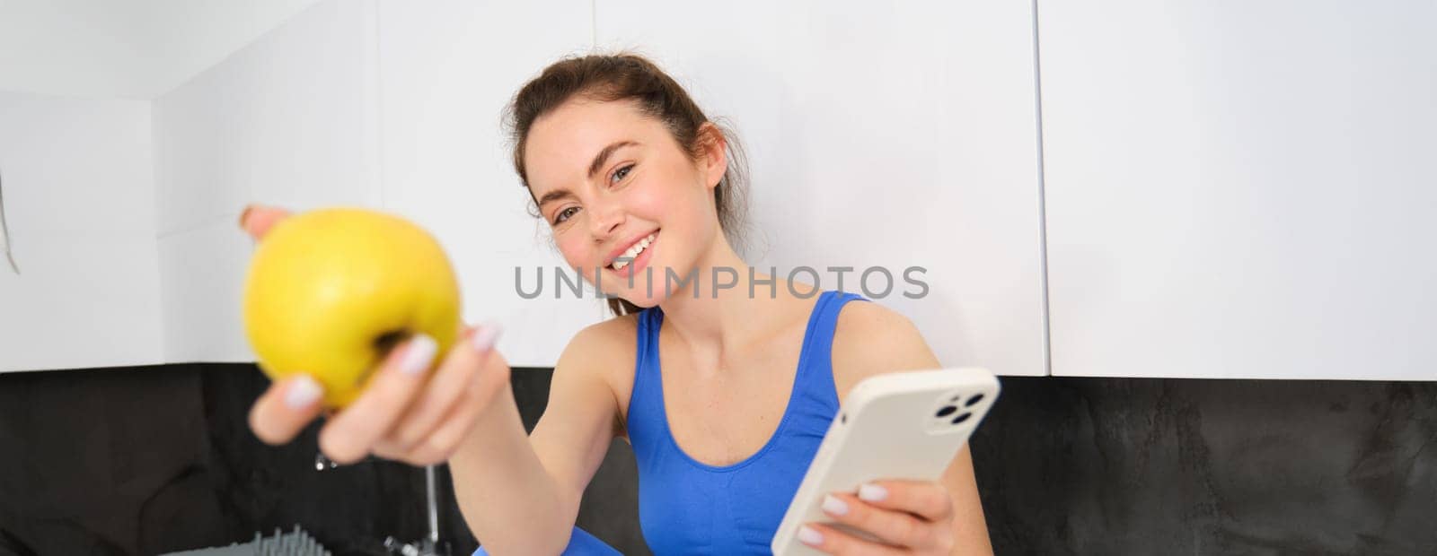Portrait of beautiful, smiling young fitness woman, offering you an apple, eating healthy snack, holding a fruit, sitting in kitchen.