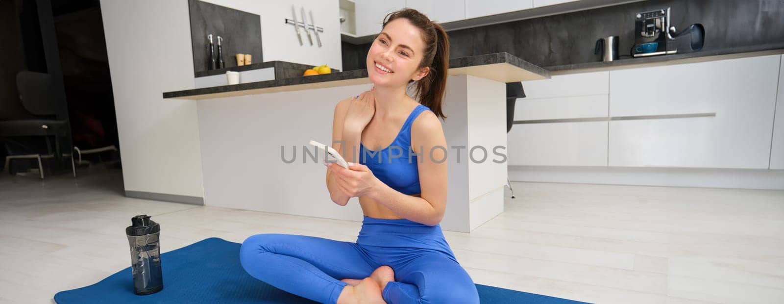 Portrait of woman using fitness app, doing workout from home, sits on yoga mat with smartphone in living room.