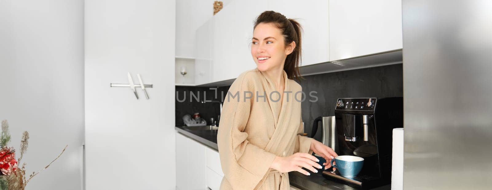 Portrait of modern woman in the morning, standing in the kitchen and making herself cup of coffee, turn around and smiling, wearing bathrobe, starting her day with delicious cappuccino.