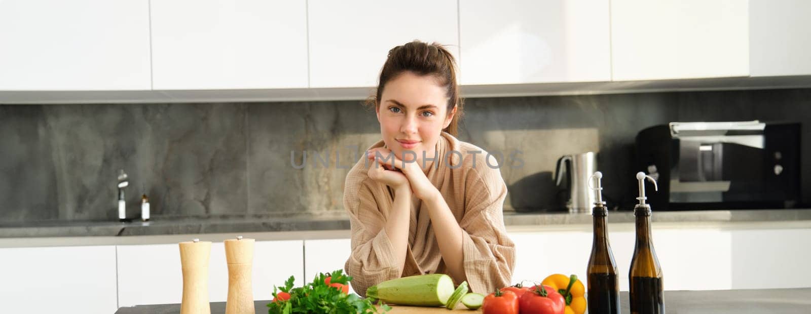 Healthy lifestyle. Young woman in bathrobe preparing food, chopping vegetables, cooking dinner on kitchen counter, standing over white background by Benzoix