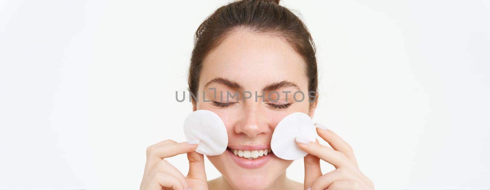 Image of young woman taking off her makeup with cotton pads, using facial cleanser, cleaning her face with skincare treatment, standing over white background.