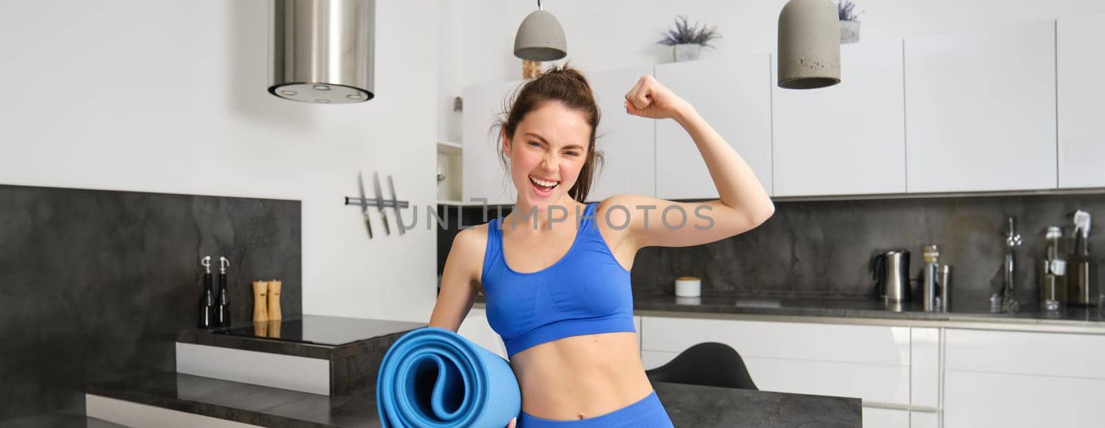 Sportswoman in track suit, shows her muscles, smiles and holds rubber yoga mat, works out at home, does sport exercises in living room.
