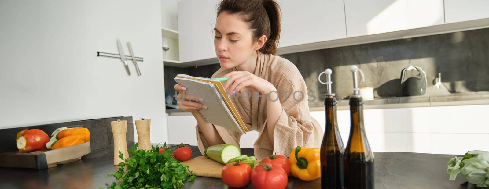 Portrait of woman checking grocery list, looking at vegetables, holding notebook, reading recipe while cooking meal in the kitchen, chopping tomatoes and zucchini by Benzoix