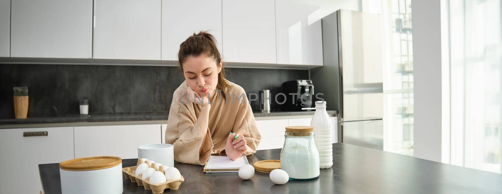 Attractive young cheerful girl baking at the kitchen, making dough, holding recipe book, having ideas.