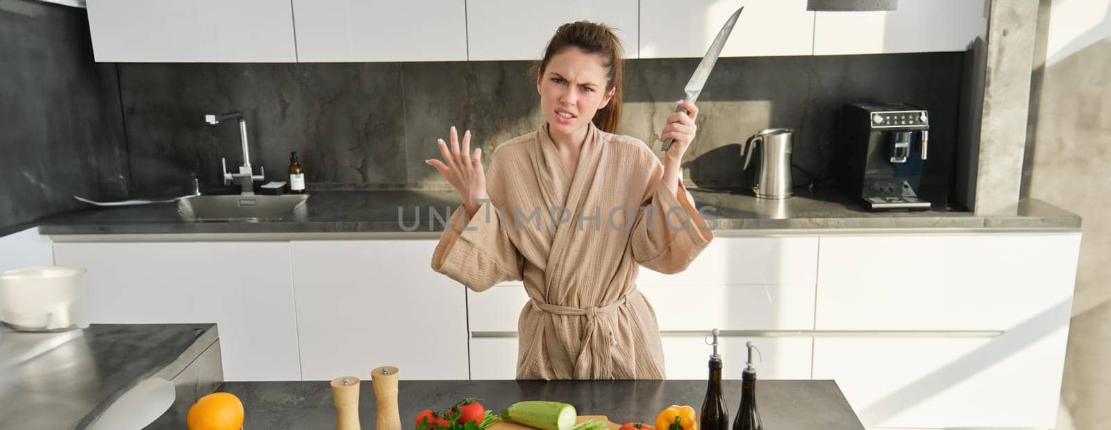 Angry brunette woman with knife, standing in the kitchen, annoyed and frustrated to cook, making upset grimace, standing in bathrobe.