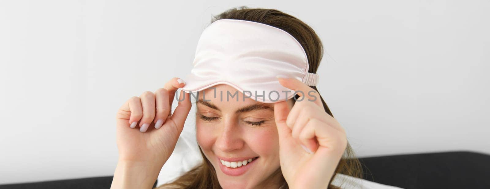 Close up portrait of gorgeous smiling woman with sleeping mask on forehead, waking up in morning, relaxing in bed, laughing and looking happy, resting on vacation in hotel room.