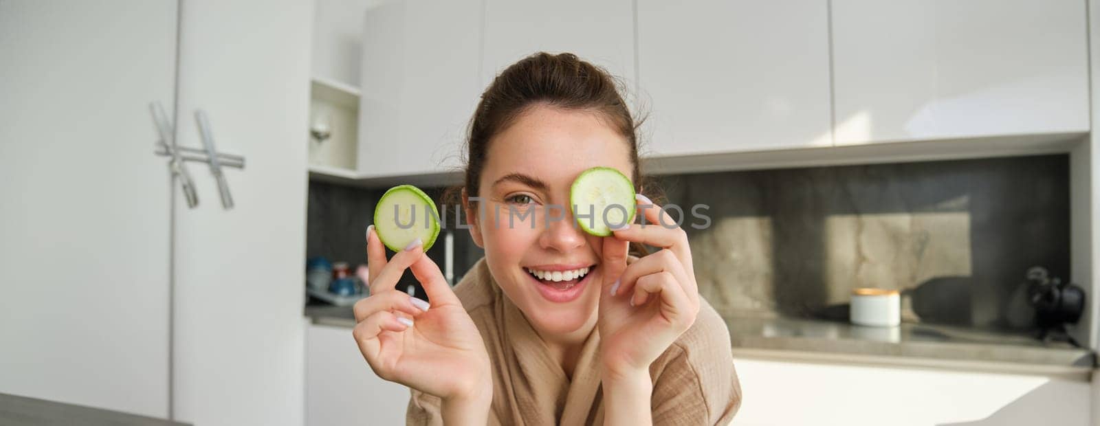 Funny, happy girl eating healthy, holding zucchini, chopping vegetables for healthy meal in the kitchen, posing in bathrobe.