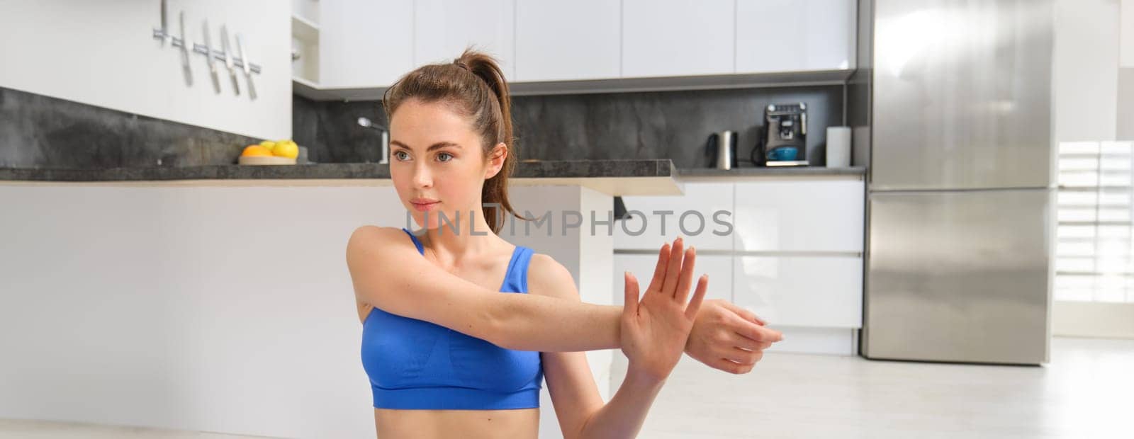 Image of fitness girl concentrates on workout, stretches hands before training session at home, follows online gym instructions by Benzoix