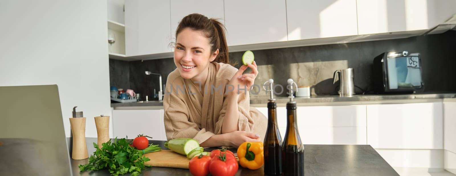 Portrait of happy girl eating vegetables while making meal, cooking healthy vegetarian food in kitchen, holding zucchini, posing in bathrobe.