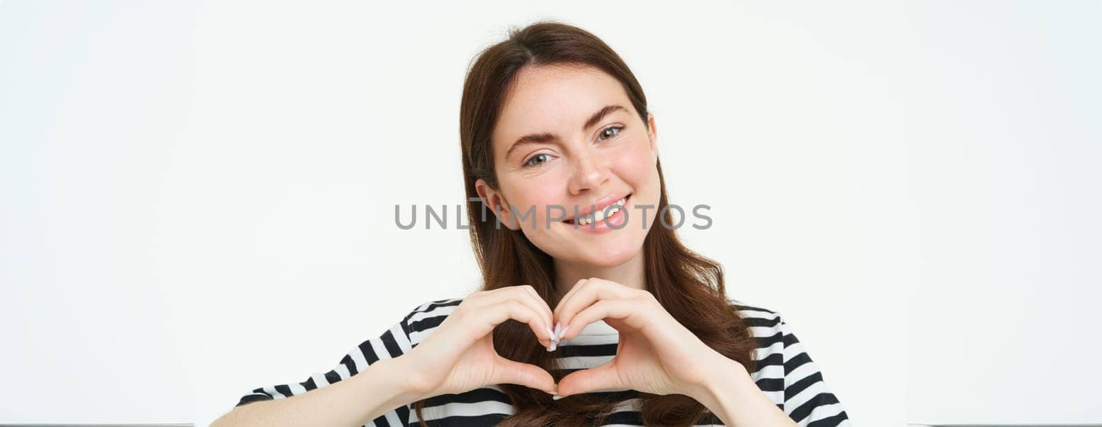 Portrait of smiling, happy girlfriend shows heart with hands and looking with care at camera, express her love or affection, like product, standing over white studio background by Benzoix