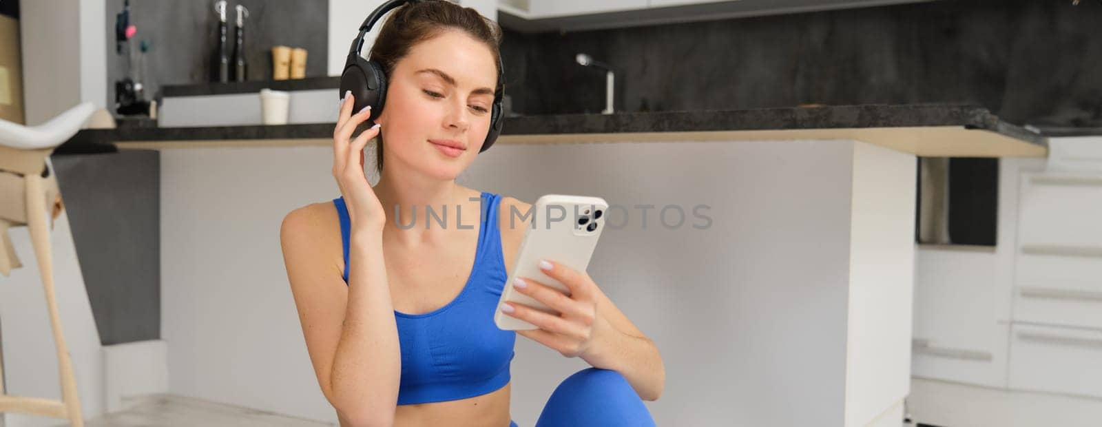 Portrait of woman in headphones, looking at smartphone, sitting on rubber mat and choosing music, doing yoga, workout training by Benzoix