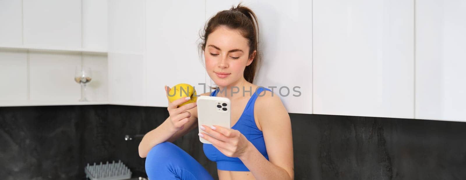 Portrait of fitness woman, sitting in kitchen with smartphone, scrolling social media and eating an apple, having a healthy snack between workout, wearing sports activewear.