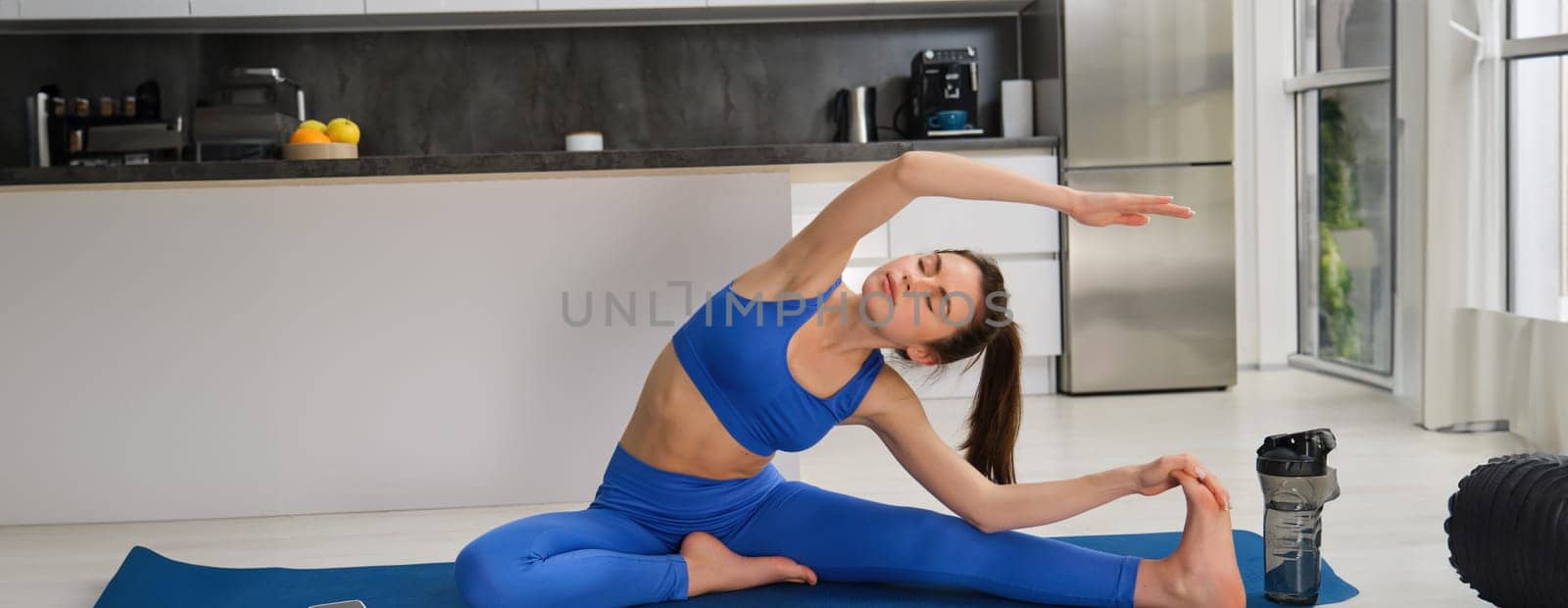 Portrait of young fitness woman doing aerobics exercises, gymnastics training at home, stretching her legs before splits workout, sitting on floor, rubber blue mat by Benzoix