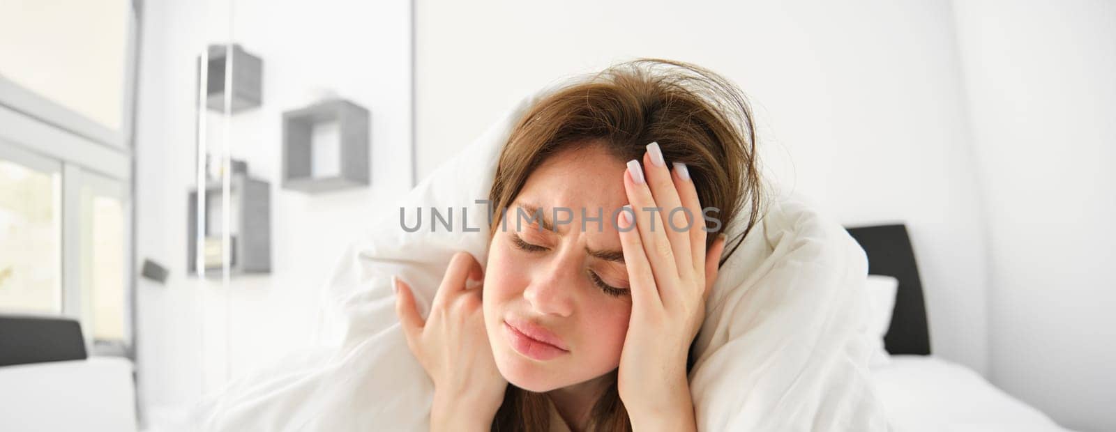 Portrait of woman with headache, lying in bed and touching head with frustrated, frowning face, does not feel well, stays in bedroom as having migraine in morning.