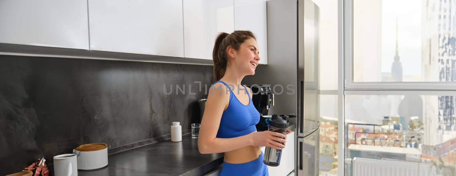 Image of stylish, beautiful young woman, finish workout, standing in kitchen at home, holding water bottle, drinkign after fitness training.