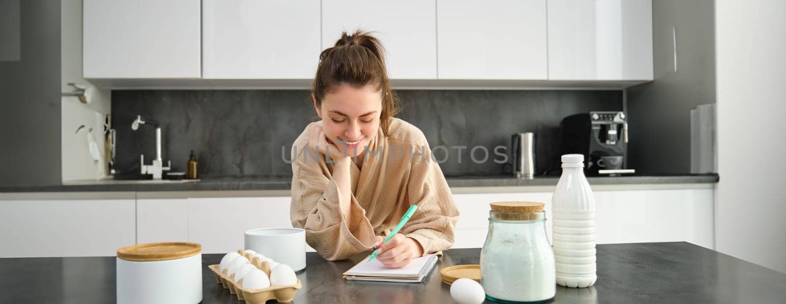 Attractive young cheerful girl baking at the kitchen, making dough, holding recipe book, having ideas.