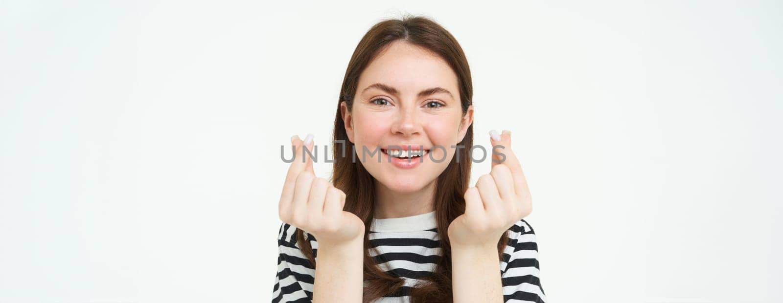 Portrait of cute young brunette woman, shows kawaii finger hearts and smiling, likes smth, stands in striped t-shirt over white background by Benzoix