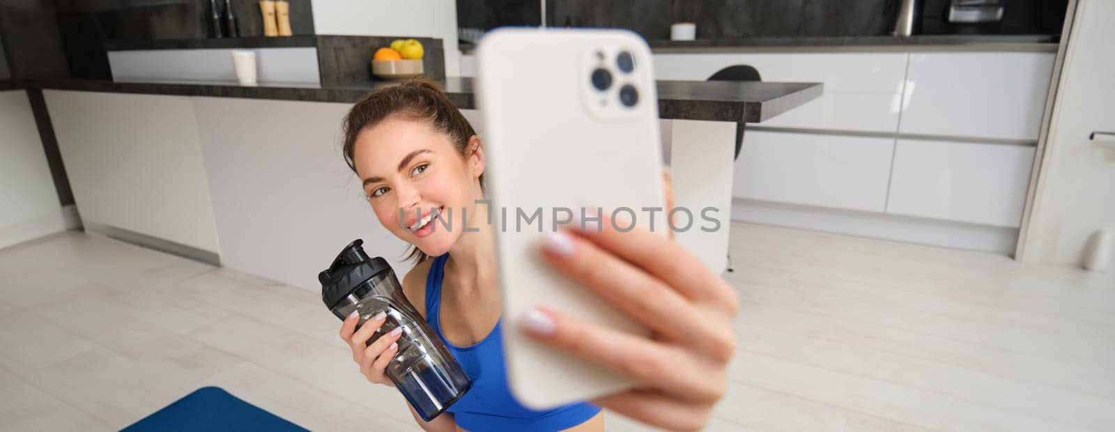 Portrait of sportswoman takes selfie with water bottle in living room, holds smartphone and poses for photo while doing fitness workout by Benzoix