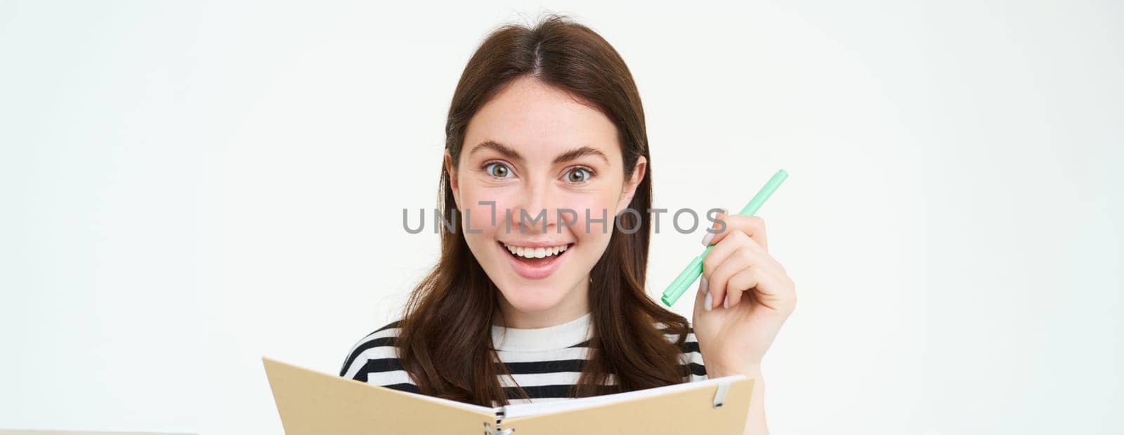 Portrait of beautiful, smiling young woman, holding pen and notebook, looking happy at camera, making notes, writing in planner, standing over white background.