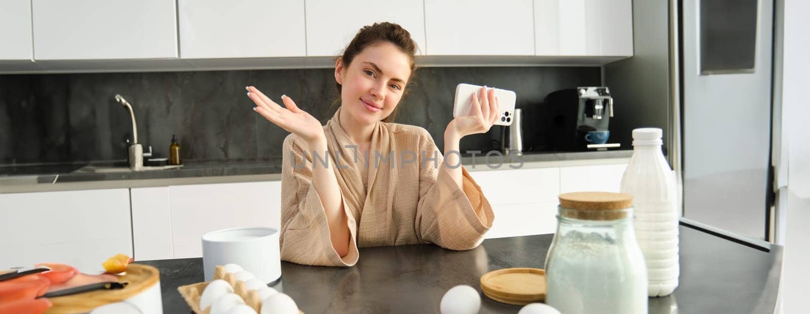 Attractive young cheerful girl baking at the kitchen, making dough, holding recipe book, having ideas.