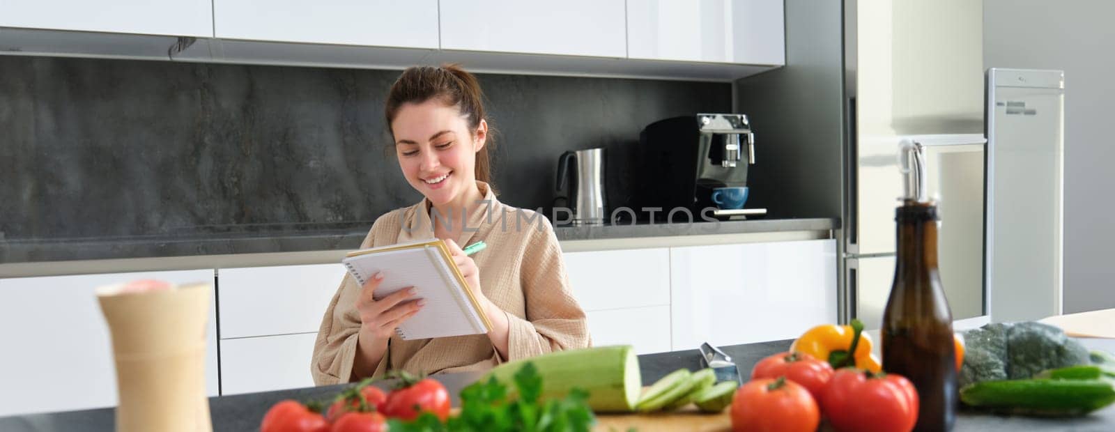 Portrait of happy young woman writes down menu for dinner, sits in the kitchen near vegetables, makes grocery list for shopping, poses in the bathrobe.