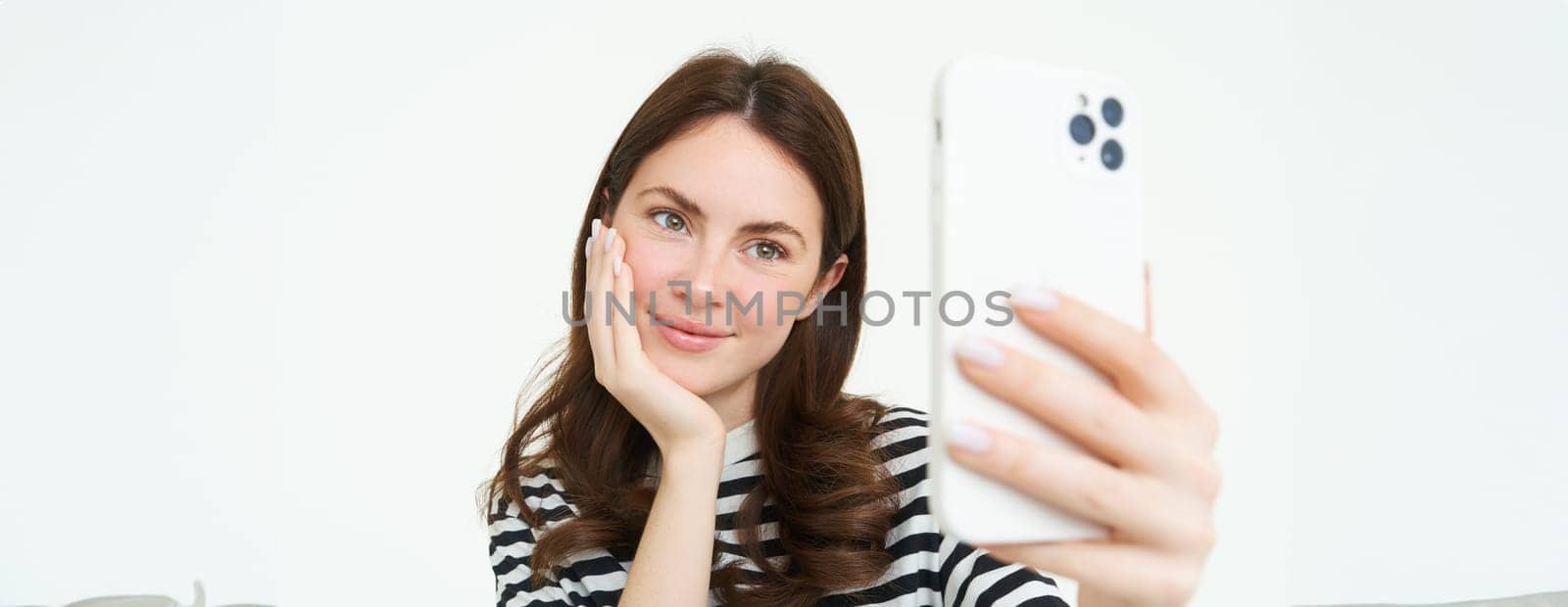 Portrait of young european woman taking selfie on smartphone, holding white mobile phone and posing for photos, isolated against white background by Benzoix