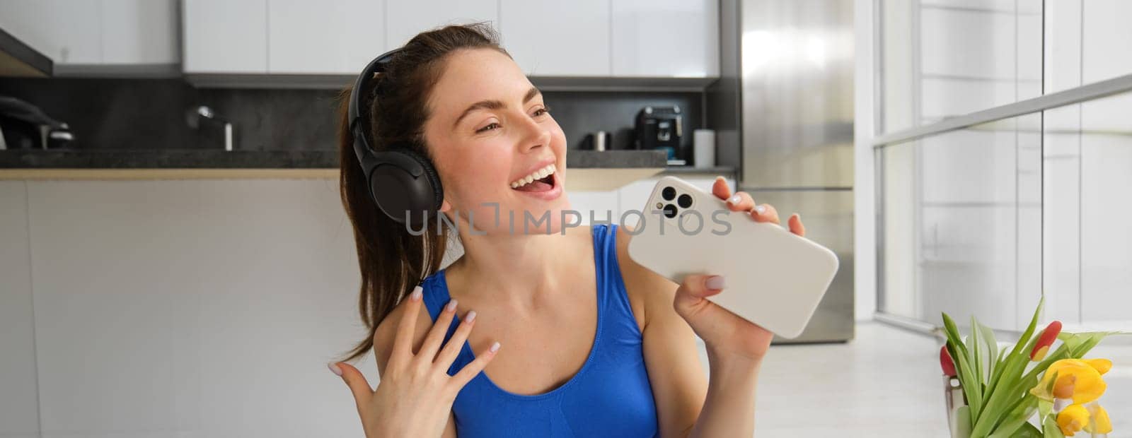 Close up portrait of happy, beautiful fitness woman, doing training exercises at home, wearing wireless headphones and singing at smartphone by Benzoix