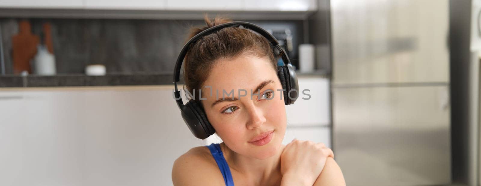 Close up portrait of brunette fitness girl, listens music in wireless headphones, smiles at camera, does workout alone at home, sits on floor and exercises by Benzoix