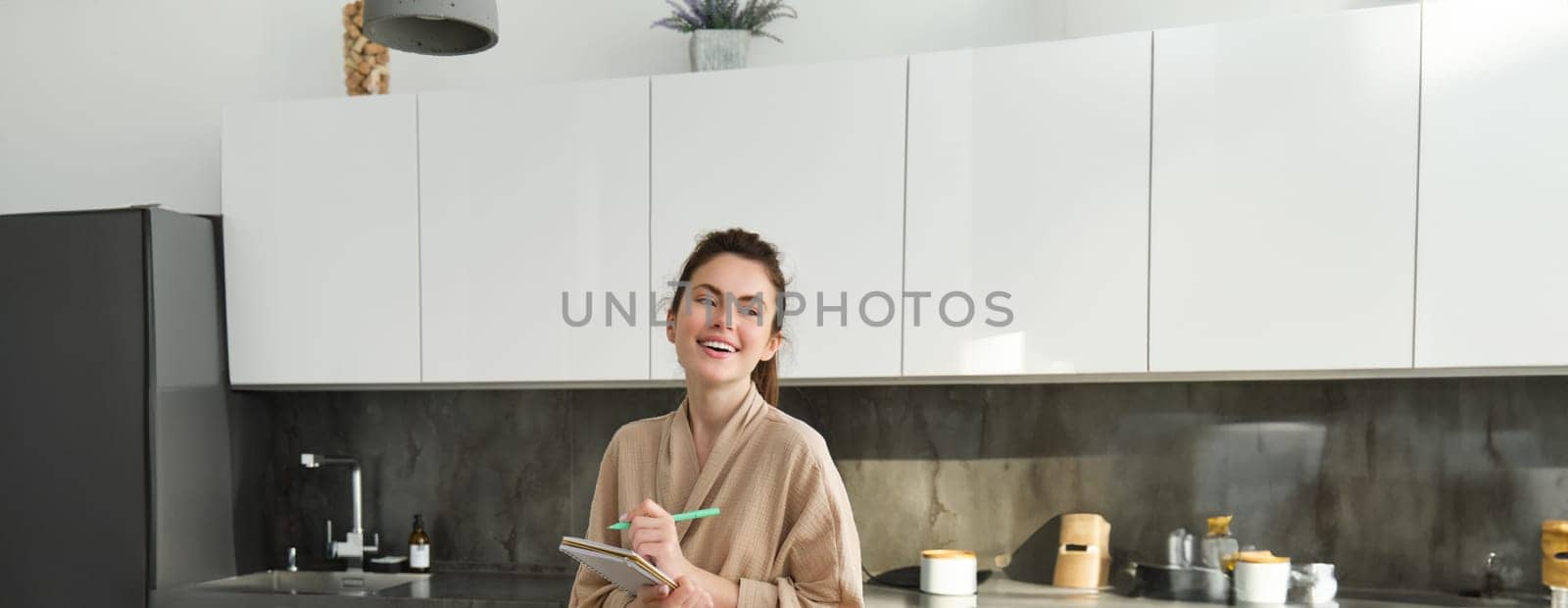 Vertical shot of young woman writing down food ideas, creating new meal, writing down grocery list or recipe in the notebook, standing in the kitchen near chopping board with fresh vegetables by Benzoix