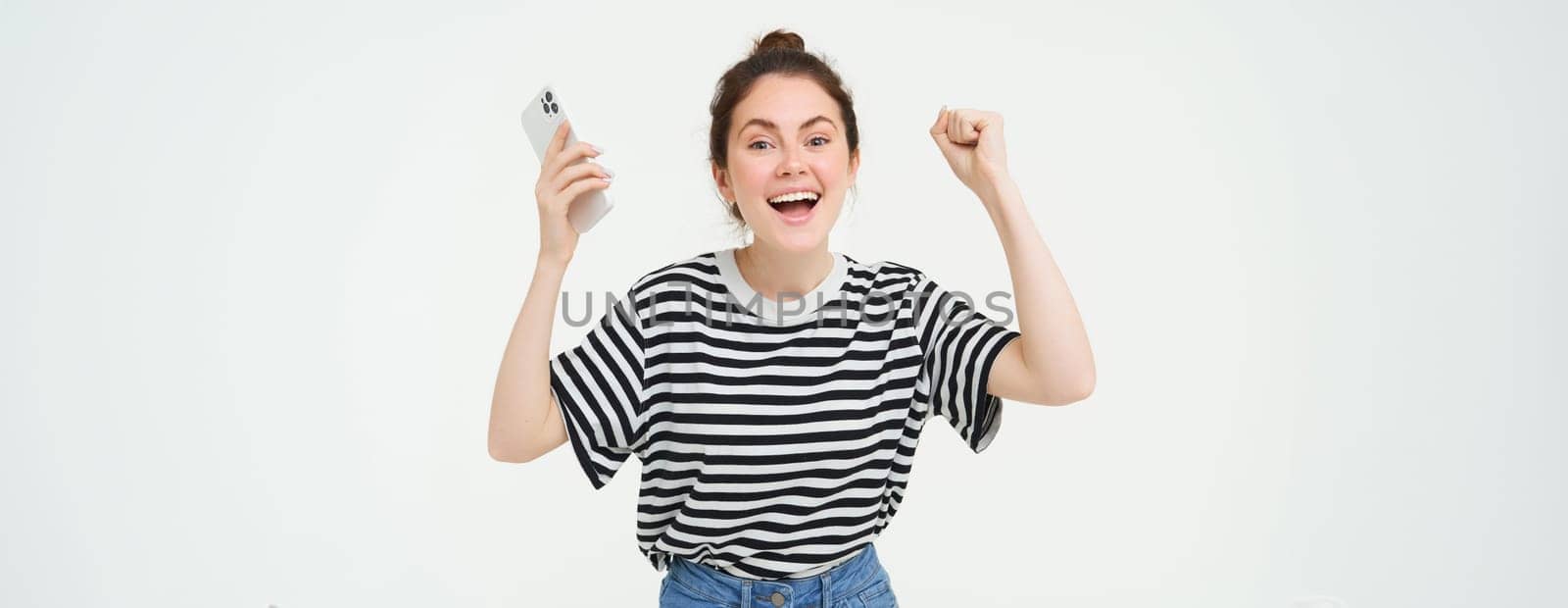 Portrait of happy woman laughing, holding telephone, using smartphone and chanting, rooting for someone, holding hands up, isolated over white background.