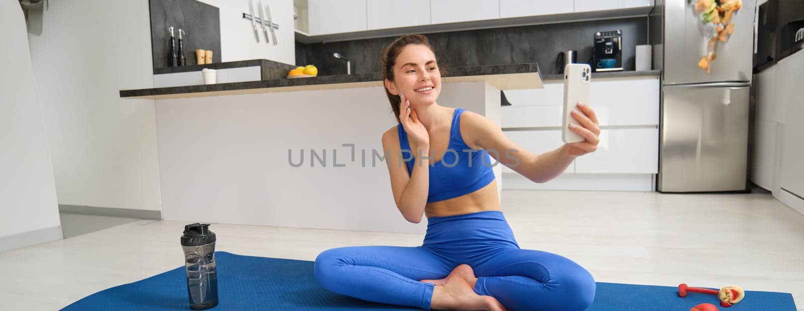 Portrait of woman workout from home, sitting on yoga mat with smartphone, taking selfie on mobile phone, doing exercises in her living room.