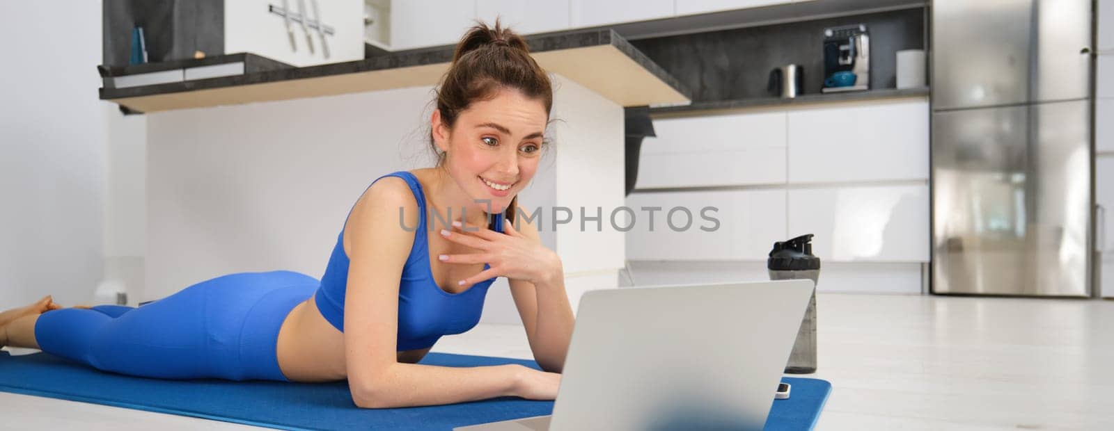 Sportswoman laying on fitness mat and looking surprised at laptop screen, talking with someone on video chat, doing workout exercises at home.