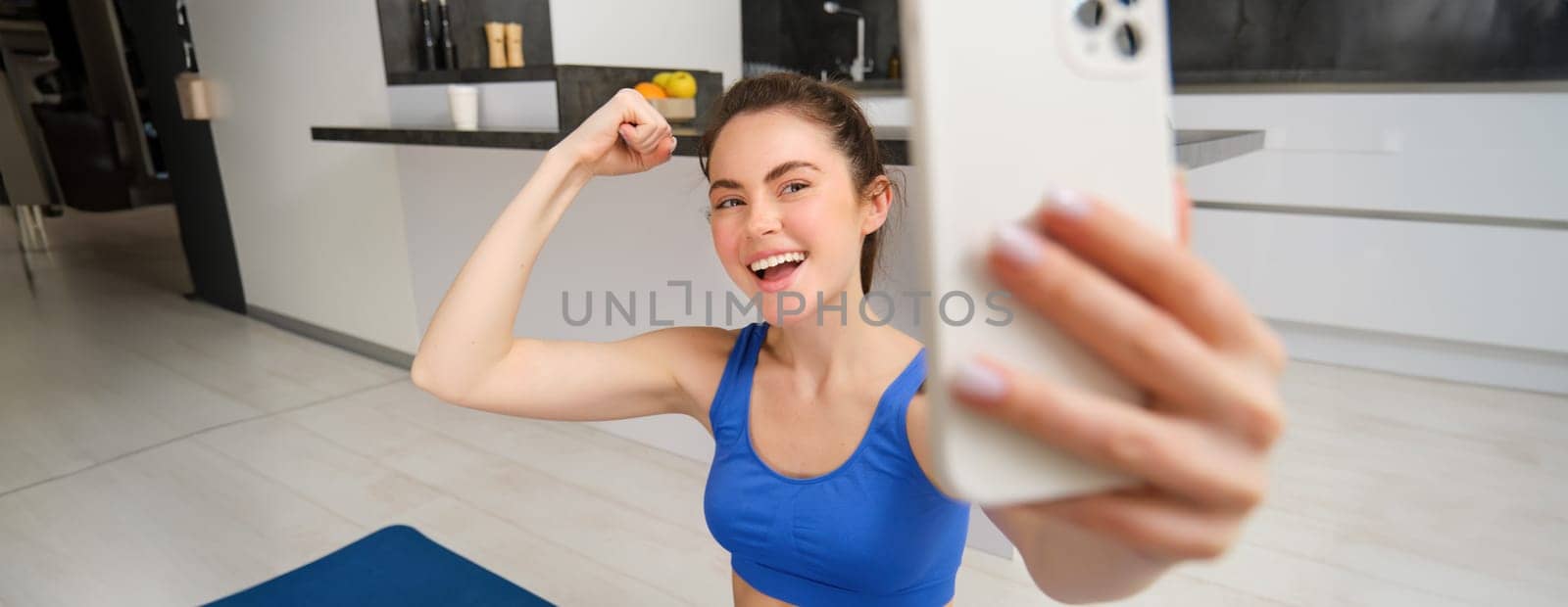 Portrait of active fitness girl, takes selfie on smartphone during home workout, posing on fitness mat, shows her strong muscles and laughs by Benzoix