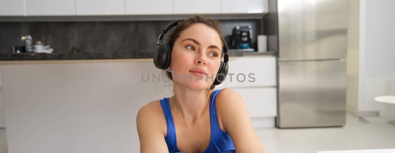 Beautiful brunette girl, doing fitness workout, sitting in living room in wireless headphones, listening to music and doing sports by Benzoix